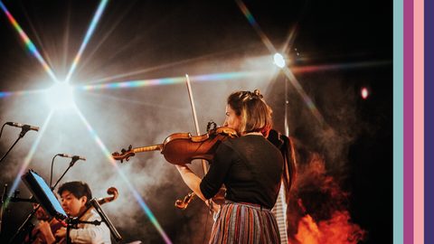 Violinist stands in foreground with back to viewer, cellist sits to the left in the background, lights are low highlighted by spotlights