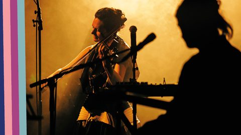 Close up of female violinist, in yellow lighting, a second person silhouetted front right