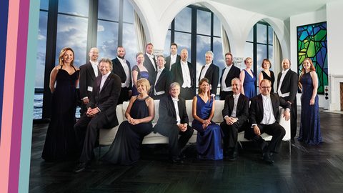 Group of people in formal dress pose for a group photo, some seated, some standing, large windows to background